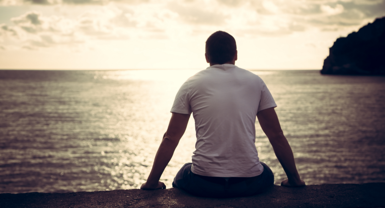 Man is sitting looking out at the sea. He has a muscular silhouette, shaved head and is slumped slightly.