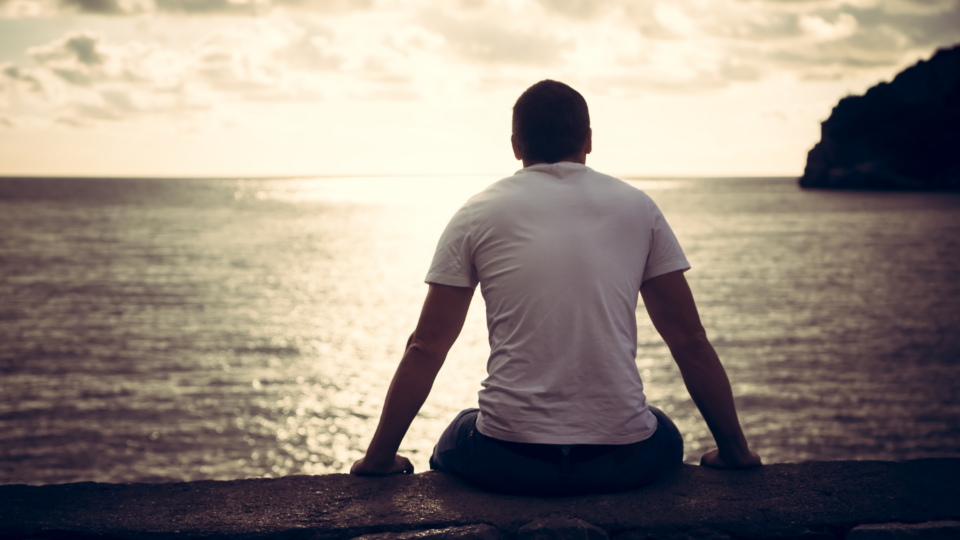 Man is sitting looking out at the sea. He has a muscular silhouette, shaved head and is slumped slightly.