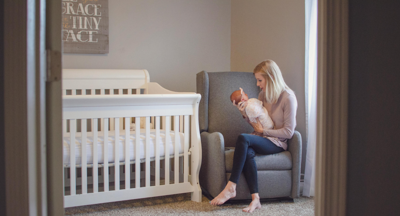 A young baby is in a swaddle next to their empty cot (safer sleep). The baby is being held by their mother who is settling the baby to sleep on their armchair.