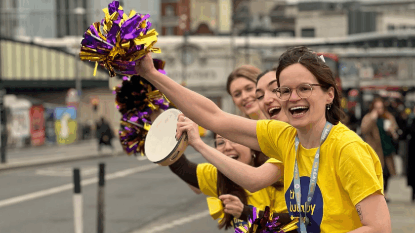 Four people in Lullaby Trust t-shirts cheer along the side of a road. The woman at the front waves a purple and gold pom-pom