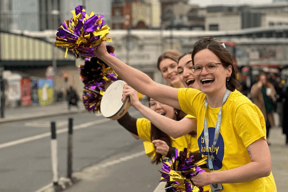 Four people in Lullaby Trust t-shirts cheer along the side of a road. The woman at the front waves a purple and gold pom-pom