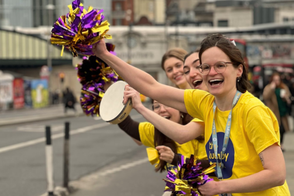 Four people in Lullaby Trust t-shirts cheer along the side of a road. The woman at the front waves a purple and gold pom-pom