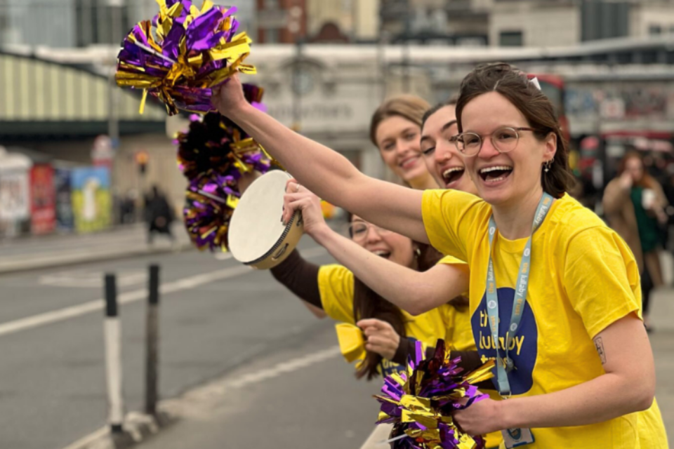 Four people in Lullaby Trust t-shirts cheer along the side of a road. The woman at the front waves a purple and gold pom-pom