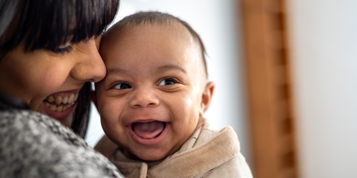 A baby is looking to their side with a huge, wide open grin. They have light to medium skin and the mum is looking down at them and smiling, with her face closely pressed to the baby.