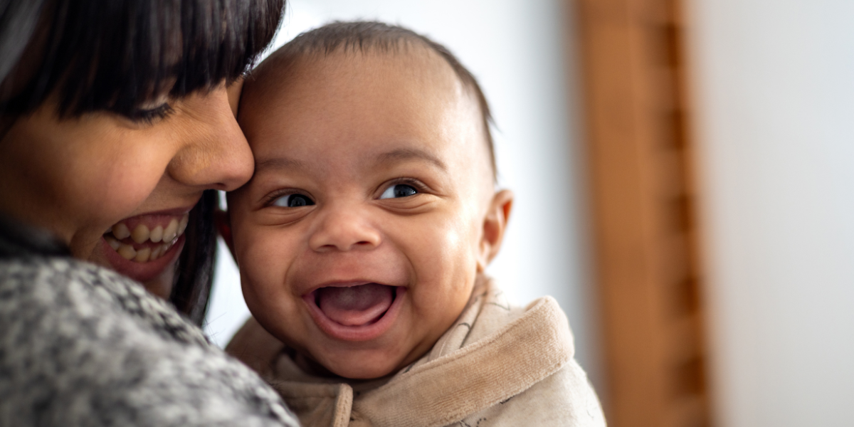 A baby is looking to their side with a huge, wide open grin. They have light to medium skin and the mum is looking down at them and smiling, with her face closely pressed to the baby.