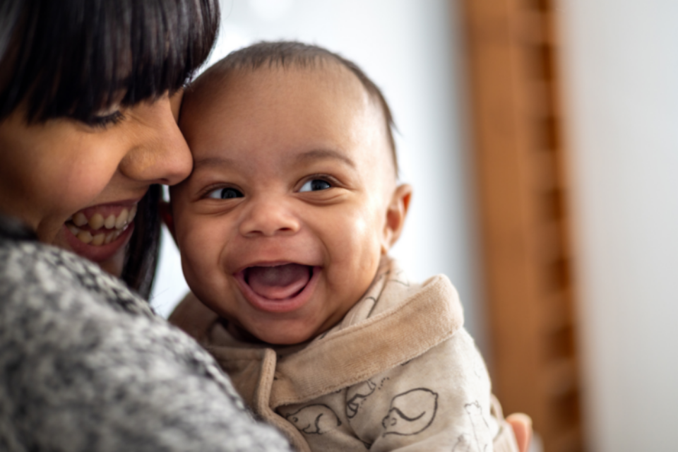 A baby is looking to their side with a huge, wide open grin. They have light to medium skin and the mum is looking down at them and smiling, with her face closely pressed to the baby.