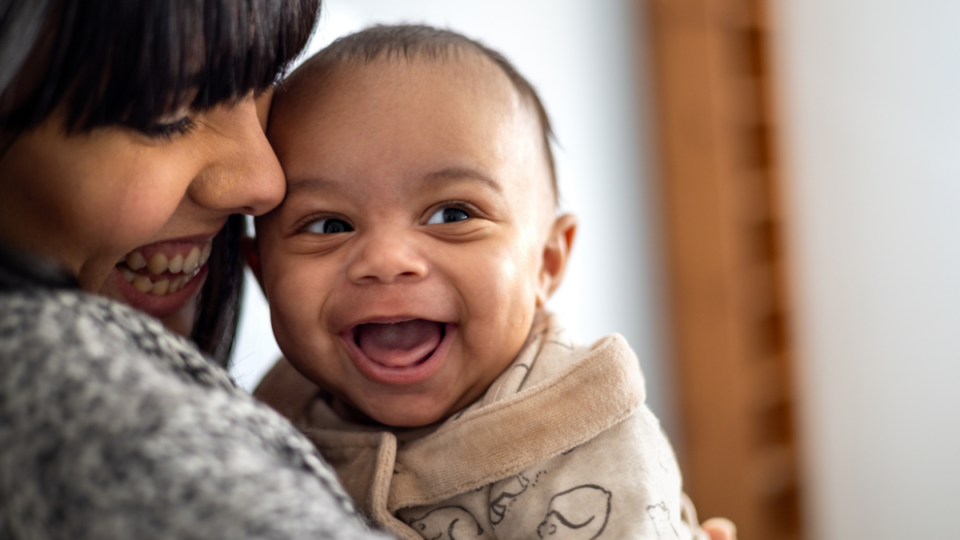 A baby is looking to their side with a huge, wide open grin. They have light to medium skin and the mum is looking down at them and smiling, with her face closely pressed to the baby.
