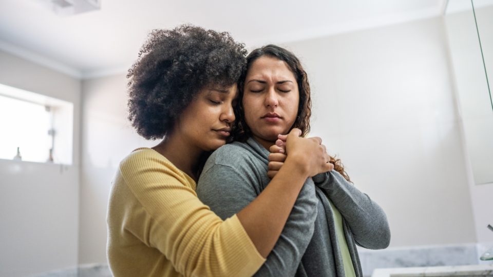 Two women are holding one another in support. They are both crying. They both have dark hair.