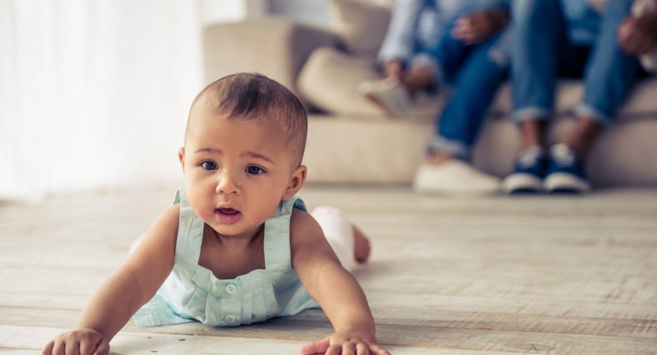 Baby is spending time on their tummy in the living room. They have brown skin and hair, and wearing a blue vest dress.
