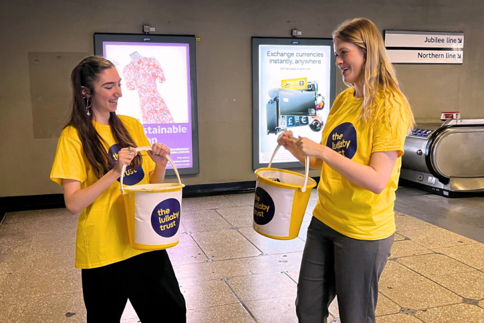 Two young women in Lullaby Trust t-shirts hold buckets to collect money in a London Underground station