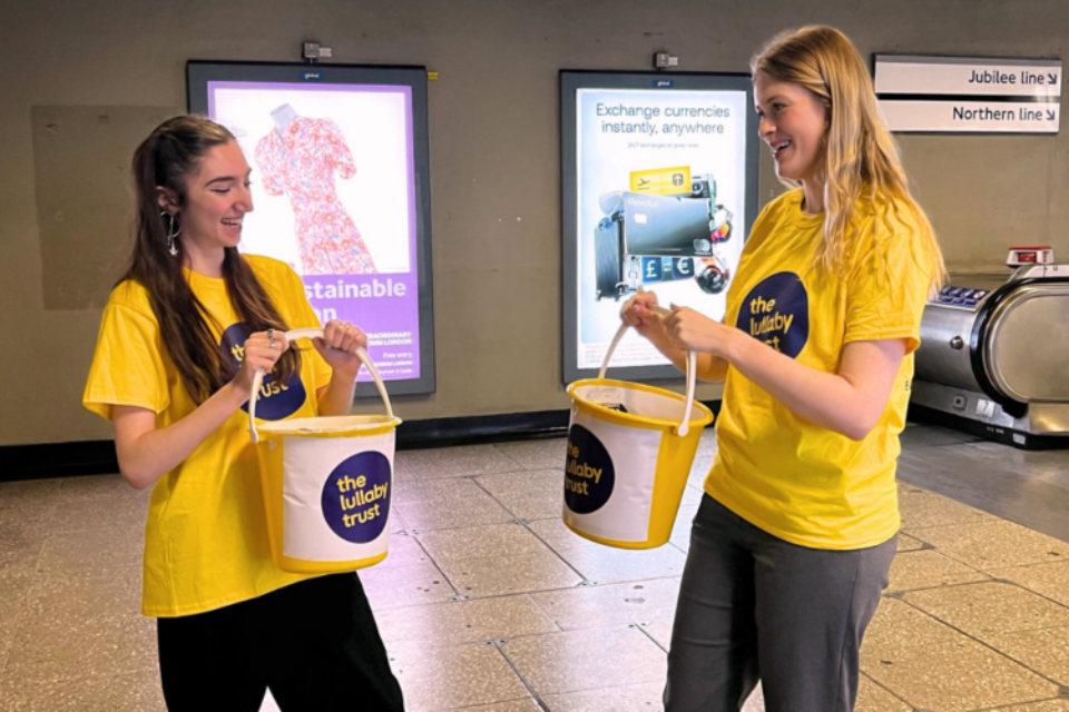 Two young women in Lullaby Trust t-shirts hold buckets to collect money in a London Underground station