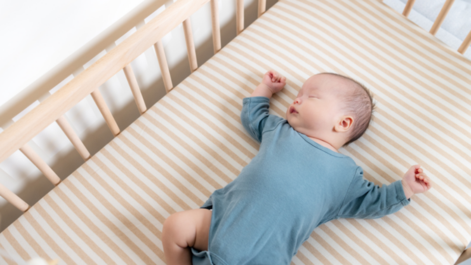 A baby in a blue baby grow is sleeping on their back in a wooden cot with a firm mattress.