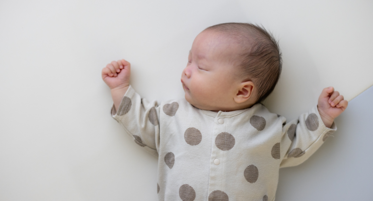 A baby is sleeping in on their back, with their head tilted to the side. They have dark hair, light skin and wear a polkadot baby grow. Their arms are rested either side of their head.