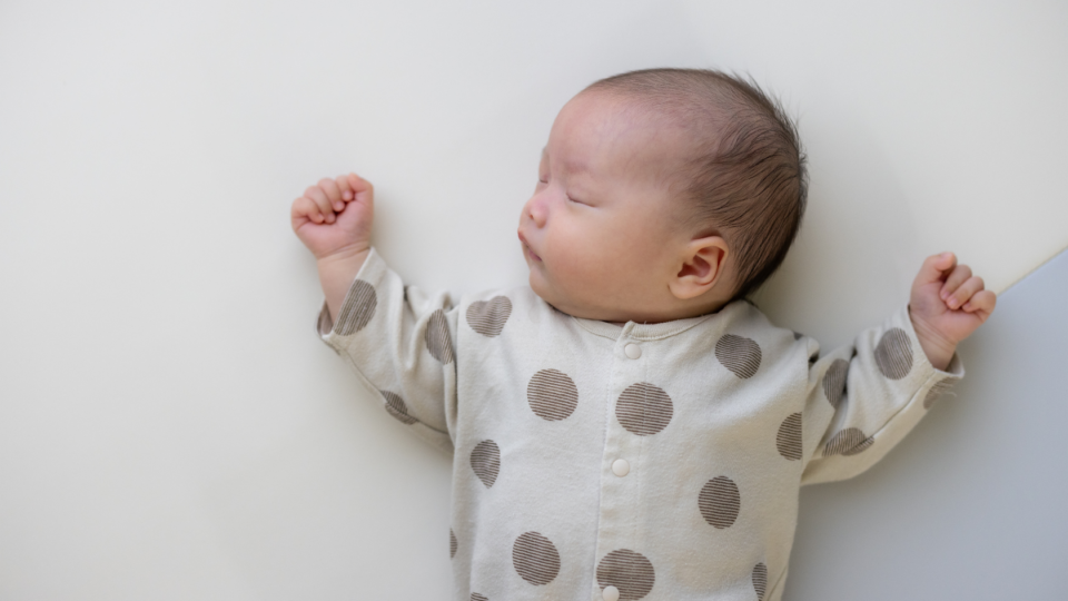 A baby is sleeping in on their back, with their head tilted to the side. They have dark hair, light skin and wear a polkadot baby grow. Their arms are rested either side of their head.