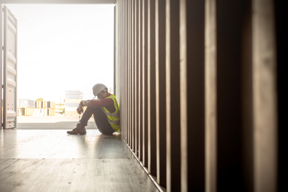 A man is slumped over in a shipping container. He is seemingly at work as wears protective gear.