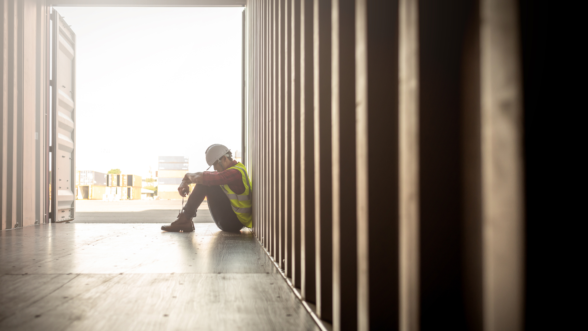 A man is slumped over in a shipping container. He is seemingly at work as wears protective gear.