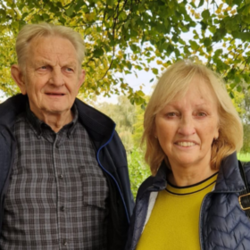 Bridget and Zbig are in a park surrounded by greenery. They are both smiling. Bridget is looking straight on, and Zbig to the side.