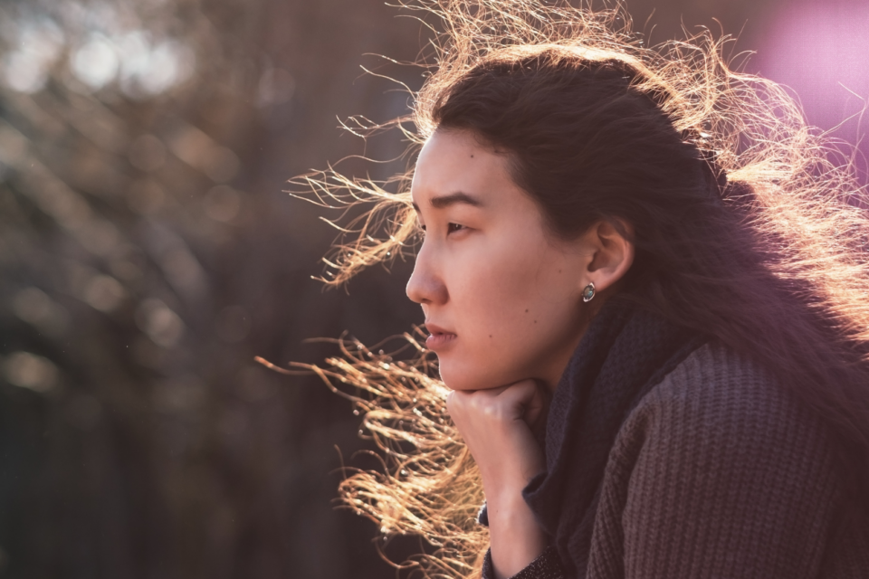 A woman is sat with an empty expression. The sun is lighting up her hair and it blows in the wind.