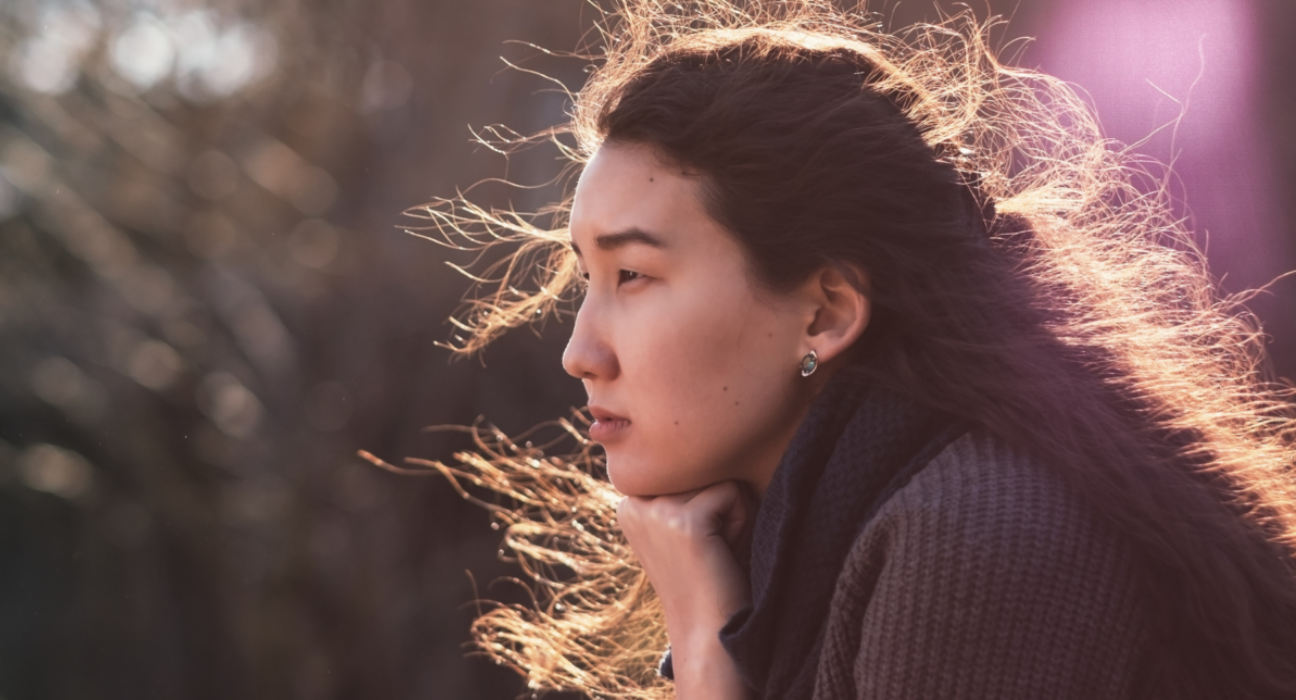 A woman is sat with an empty expression. She is looking out, and the sun is behind her dark hair which is blowing in the wind.