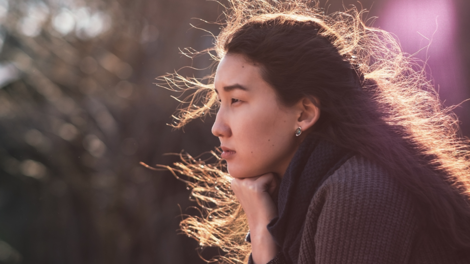 A woman is sat with an empty expression. The sun is lighting up her hair and it blows in the wind.