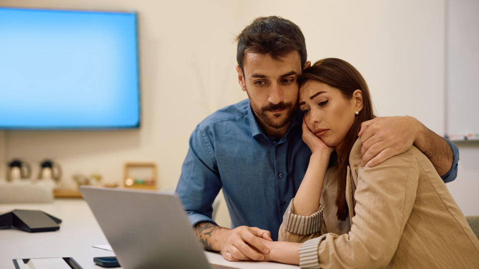 A couple are sat looking at their laptop, leaning on one another. They look worried.