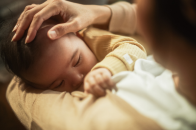 A baby is breastfeeding. They are clutching onto their mother's t-shirt and wear a light yellow top. The mum has the hand gently placed on the baby's head.