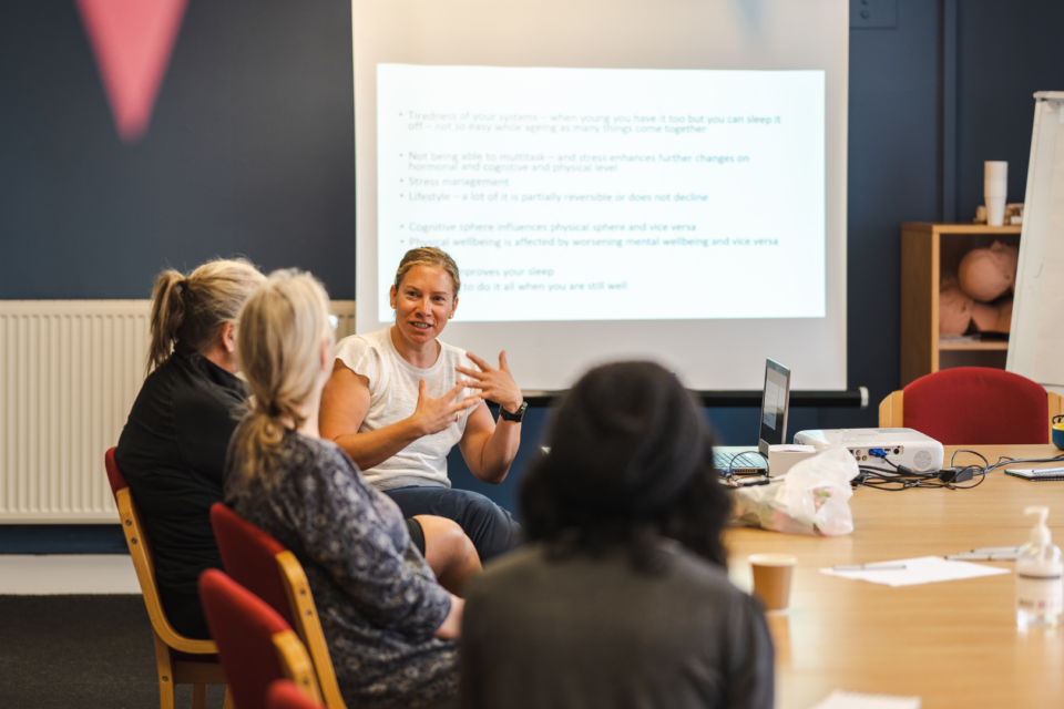 A group of people are at a meeting. A woman is speaking to another three or four people on one side of a table, there is a presentation behind her.