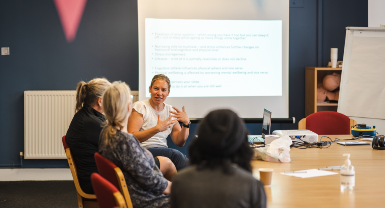 A group of people are at a meeting. A woman is speaking to another three or four people on one side of a table, there is a presentation behind her.