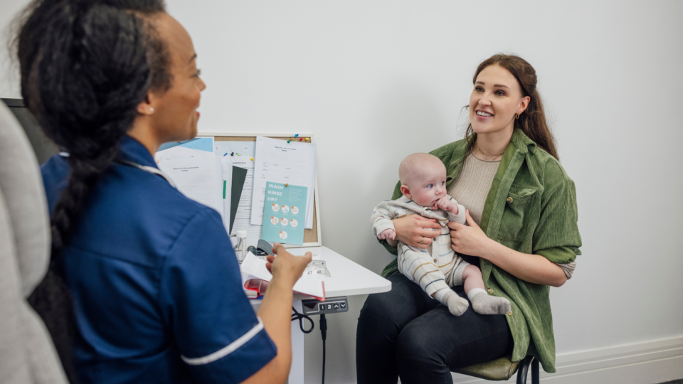 Mother sitting on a chair in a doctor's office with a baby on her lap, talking to a health professional.