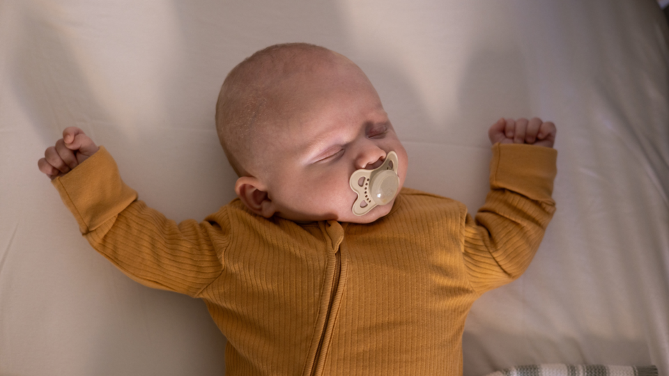A baby is asleep in a safer sleep position, on their back with a clear cot. They have a dummy in their mouth