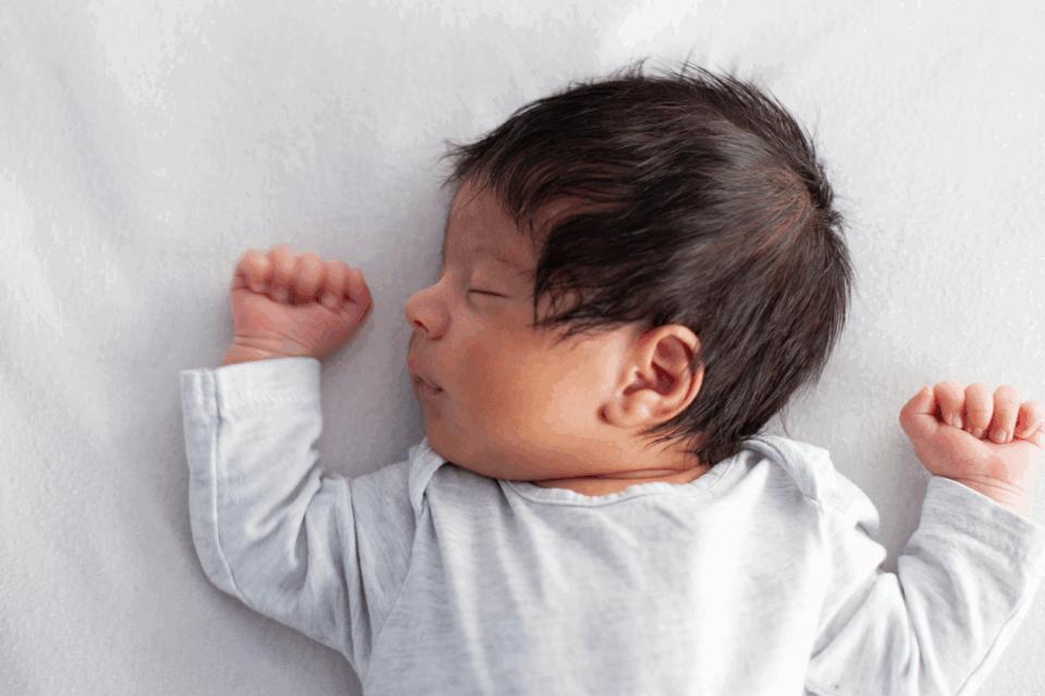 A baby is sleeping in a safer sleep position. They are on their back with a clear cot around them. They have dark hair and medium skin, and their arms are held up at either side of their head. They are sleeping.