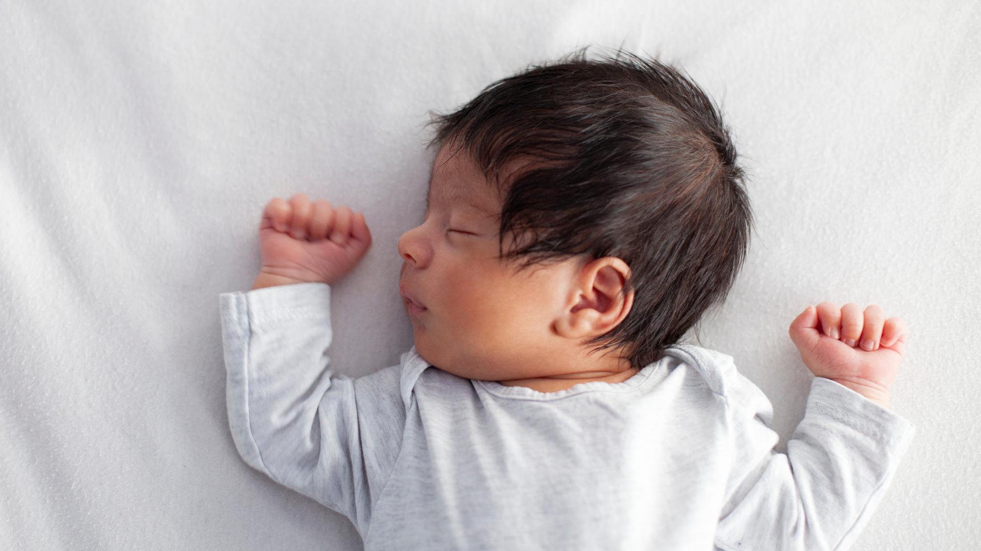 A baby is sleeping in a safer sleep position. They are on their back with a clear cot around them. They have dark hair and medium skin, and their arms are held up at either side of their head. They are sleeping.