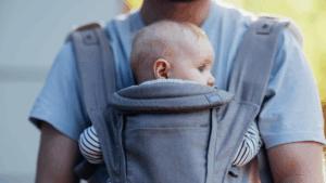 A baby is in a baby carrier attached to their dad's chest. They are looking around. It is a safer position for baby as their airway is clear and dad could kiss baby's head if he tried.