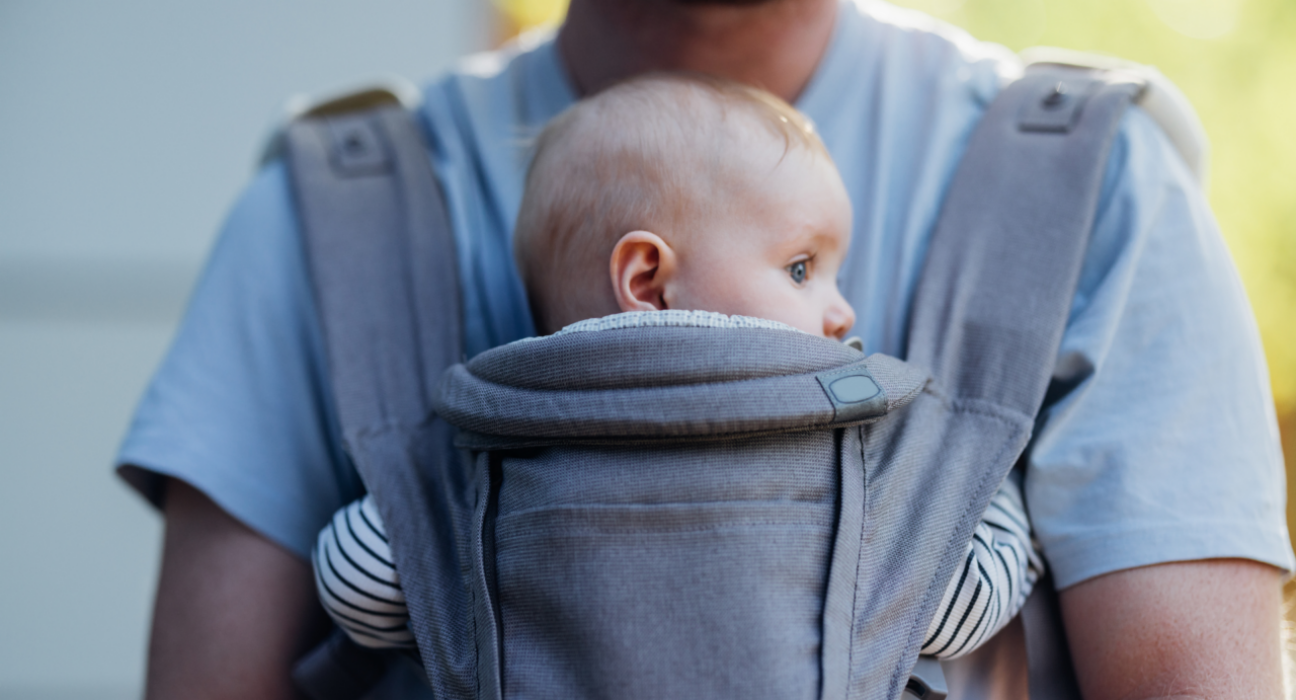 A baby is in a baby carrier attached to their dad's chest. They are looking around. It is a safer position for baby as their airway is clear and dad could kiss baby's head if he tried.