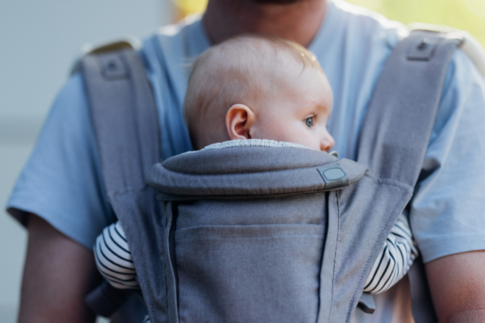 A baby is in a baby carrier attached to their dad's chest. They are looking around. It is a safer position for baby as their airway is clear and dad could kiss baby's head if he tried.