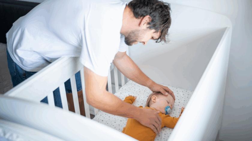 A baby is sleeping in a safer sleep position in a cot. The dad is holding a hand on his chest and head. The baby has a dummy.