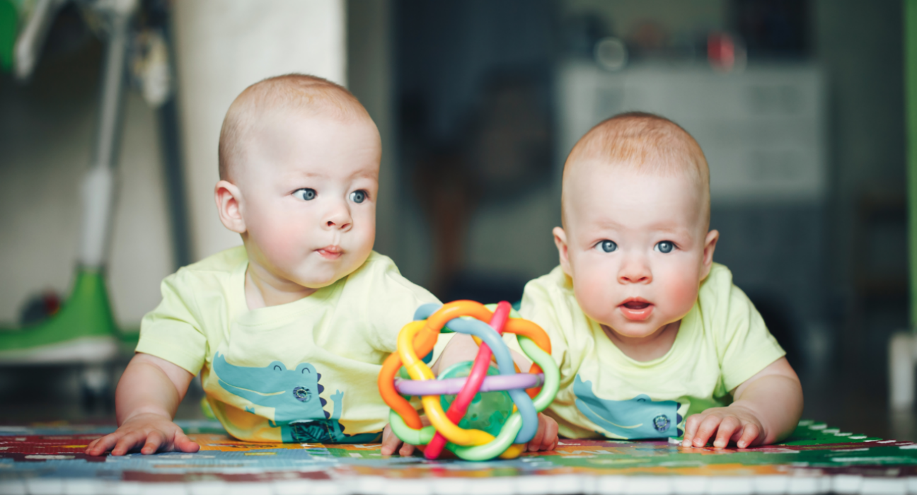 Two young twin babies are having tummy time on a play mat with a toy. They have ginger hair and light skin with bright blue eyes.