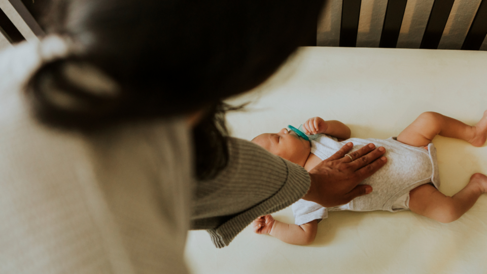 A baby is sleeping on their back in their cot with a dummy in their mouth. They have a medium skin tone and their mum has her hand on their chest.
