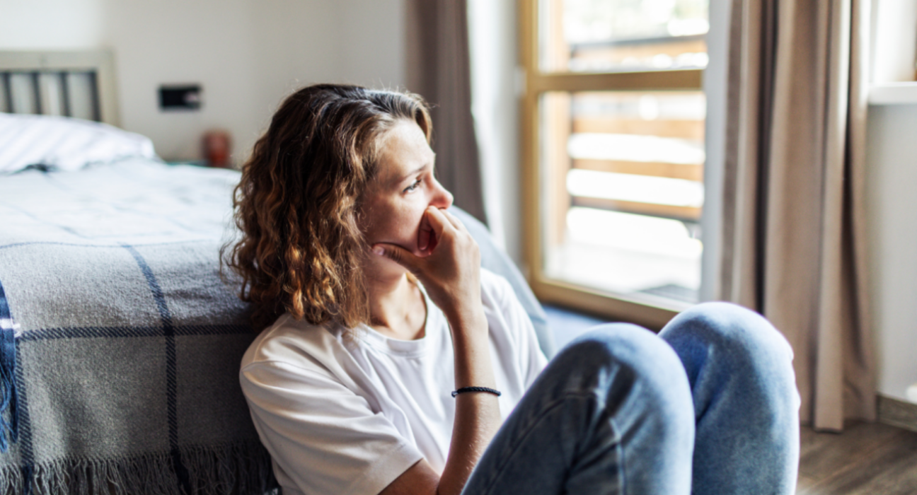 Lady sitting on the floor at the end of her bed, looking out towards the window sadly.