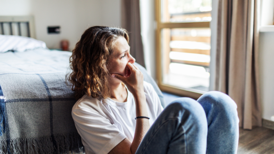 Lady sitting on the floor at the end of her bed, looking out towards the window sadly.
