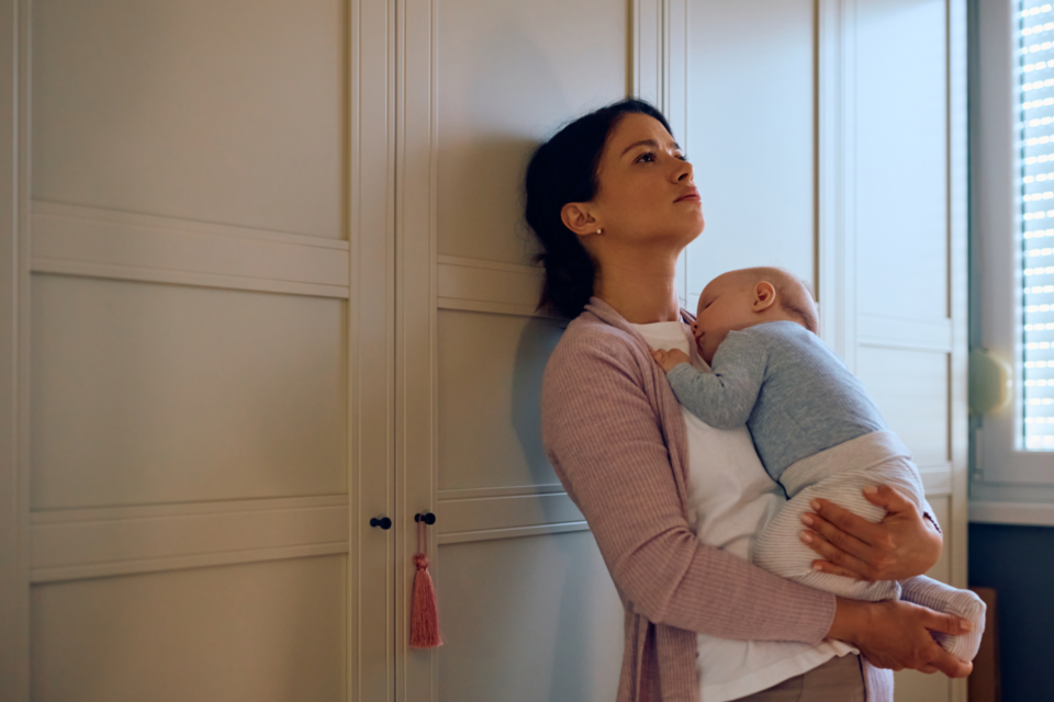 Lady standing, holding her baby and leaning against a wardrobe.