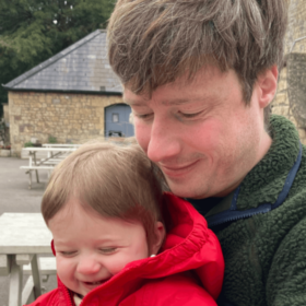 Matt and baby Sam are sat on a bench. Sam is grinning cheekily and Matt is holding him, smiling and looking down at baby Sam's hands.