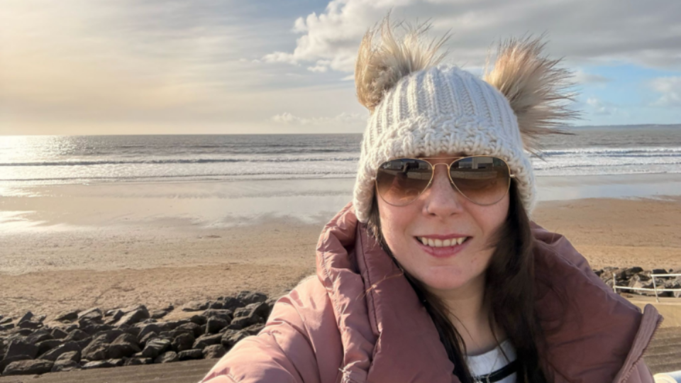 Becky taking a selfie by the sea, wearing a wooly hat and smiling at the camera.