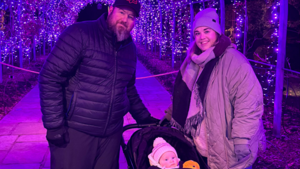 Camila and James standing next to Sophia in her buggy, surrounded by festive lights.