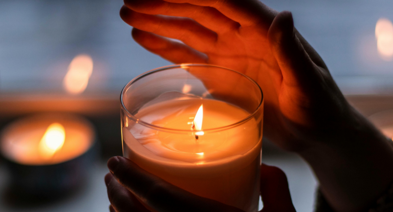 A person has lit a candle and they are holding it in one hand, and holding their other hand above the candle which is in a glass jar. In the blurred background, you can see another candle on a windowsil.