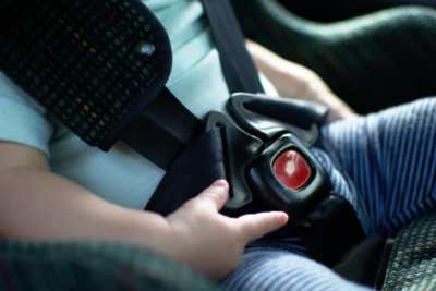 Close up of a baby in a car seat. You can see their mouth and the car seat buckled across their blue t-shirt. They are playing with the plastic around the buckle.