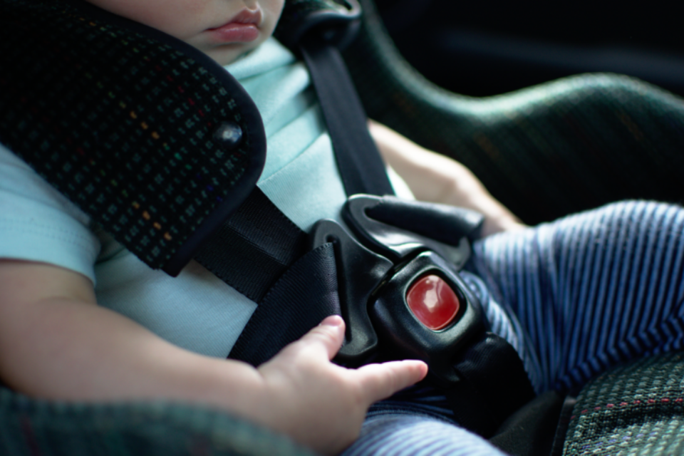 Close up of a baby in a car seat. You can see their mouth and the car seat buckled across their blue t-shirt. They are playing with the plastic around the buckle.