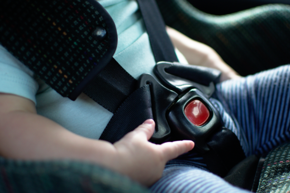 Close up of a baby in a car seat. You can see their mouth and the car seat buckled across their blue t-shirt. They are playing with the plastic around the buckle.