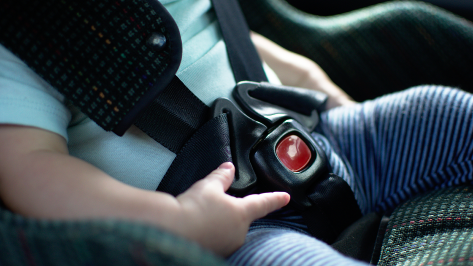 Close up of a baby in a car seat. You can see their mouth and the car seat buckled across their blue t-shirt. They are playing with the plastic around the buckle.