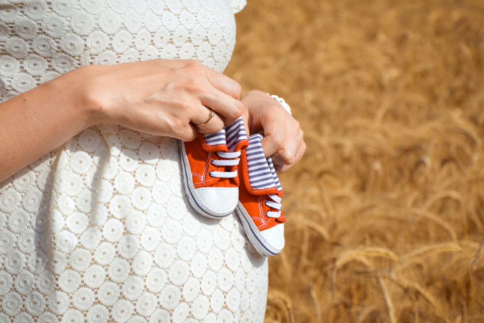 Person holding baby's shoes in front of her bump, wearing a white dress.